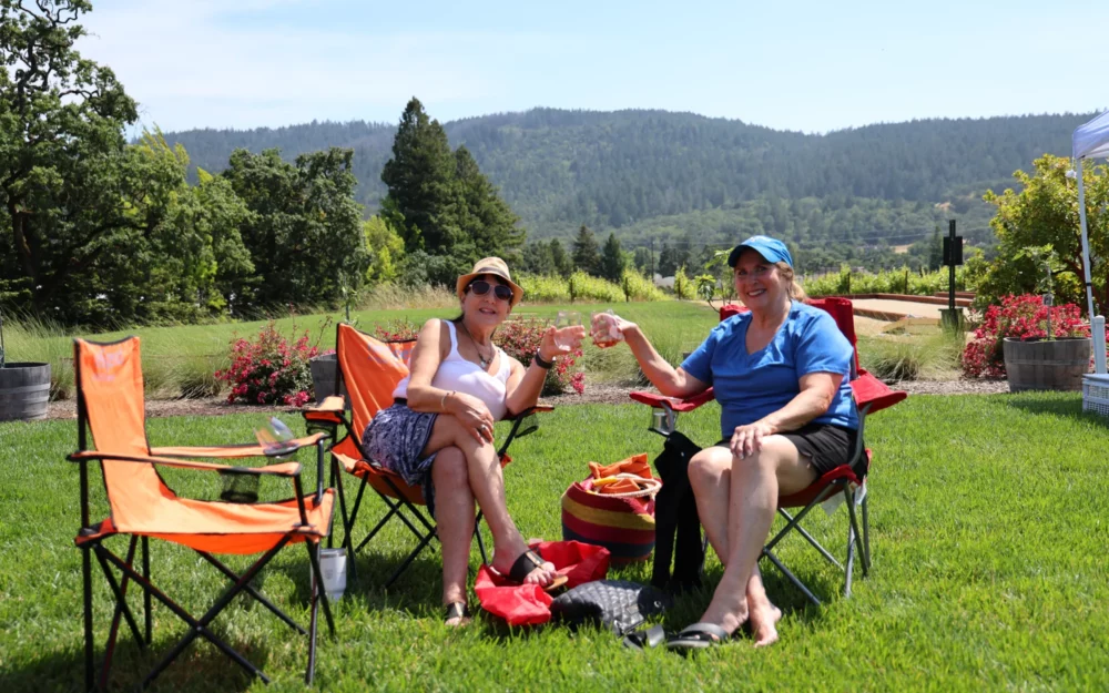 Two women in lawn chairs toasting with wine glasses at Sugarloaf Wine Co., winery lawns, outdoor event venue with live music, Santa Rosa, CA,